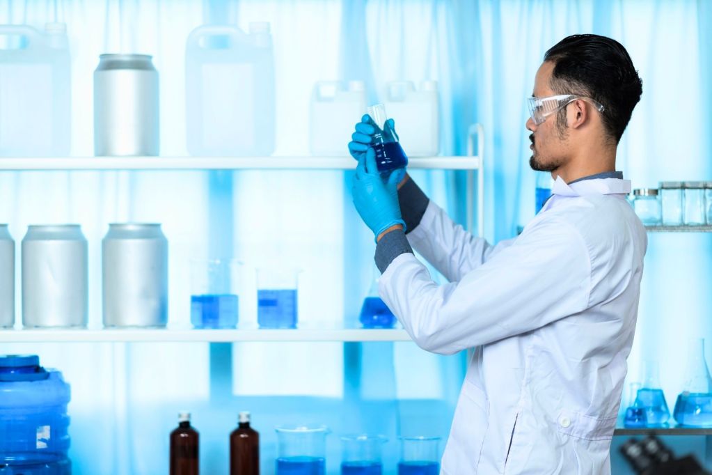 A man in a white lab coat and wearing safety glasses and blue gloves looks at a piece of glassware filled with liquid
