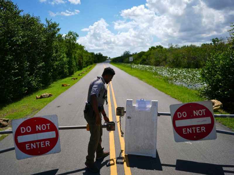 A man closes a gate on a road. A “Do Not Enter” sign hangs on each side of the gate.