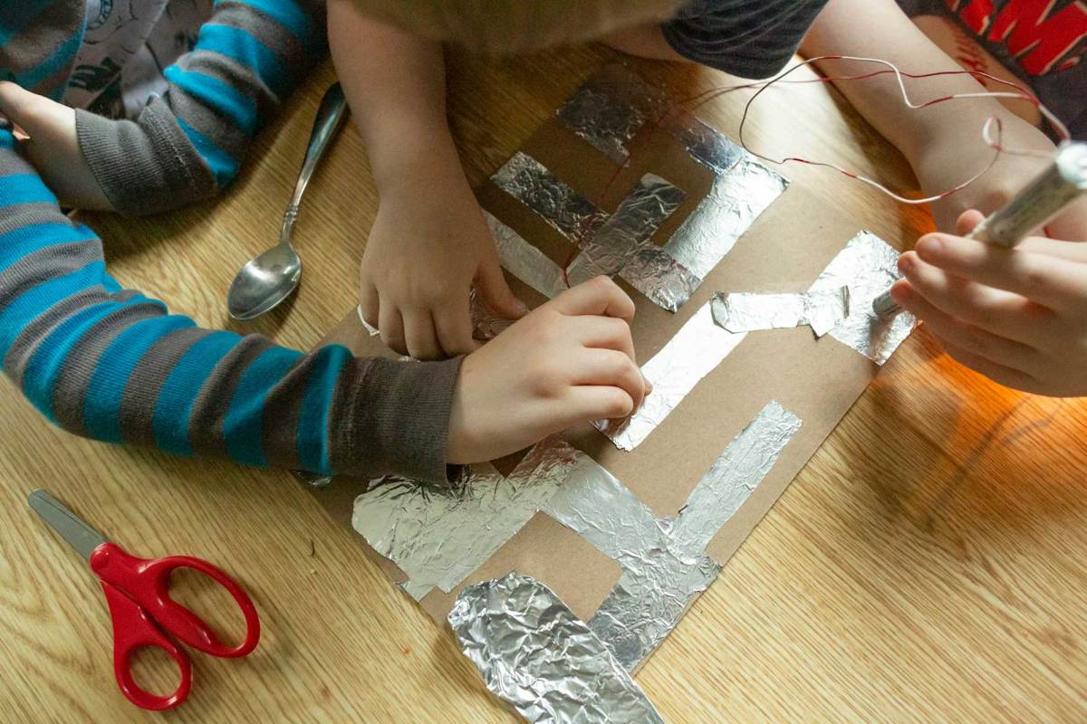 An art project made of cardboard and aluminum foil on a table, with hands from different people working on it.