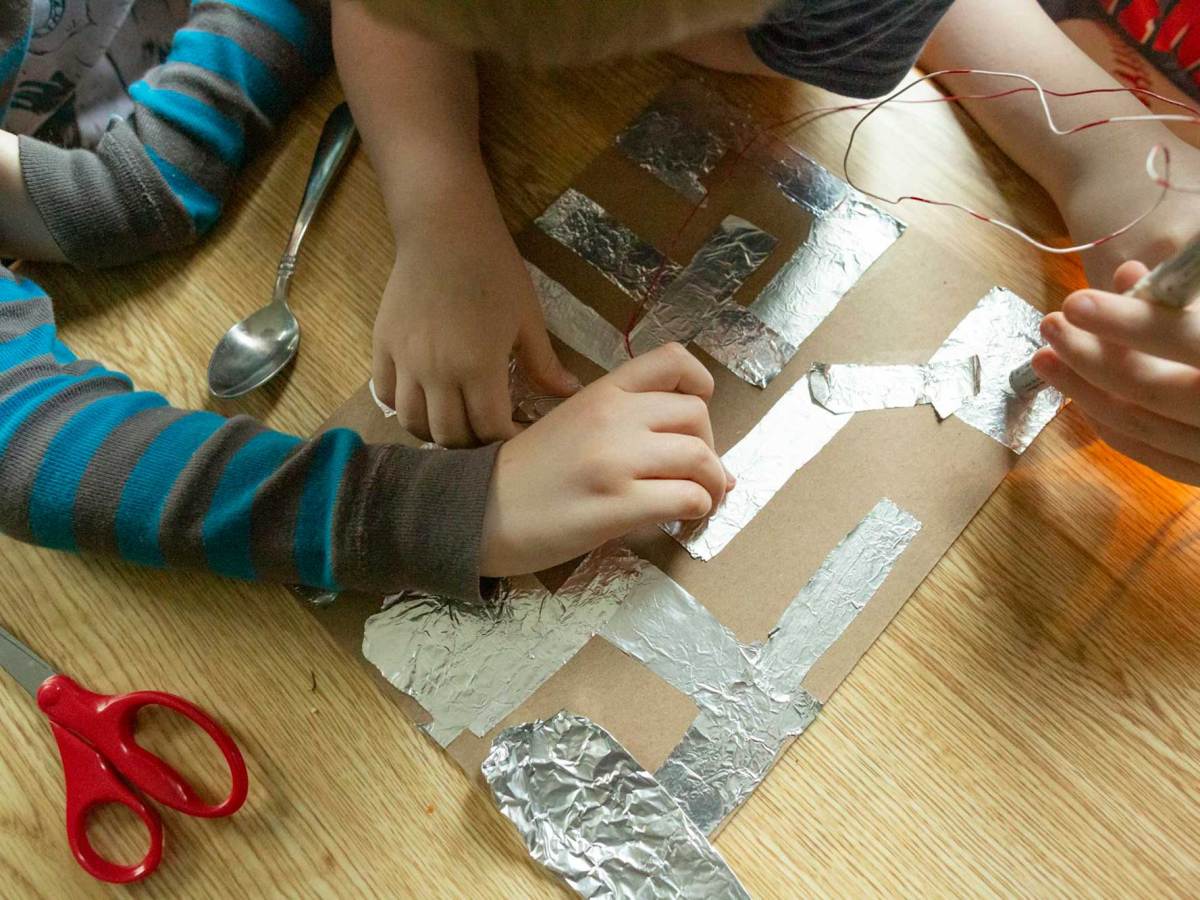 An art project made of cardboard and aluminum foil on a table, with hands from different people working on it.