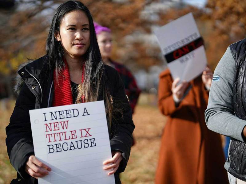 A photo of a dark-haired woman at a protest holding a sign that reads ‘I need a new title ix because…’