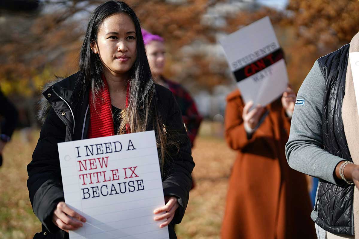 A photo of a dark-haired woman at a protest holding a sign that reads ‘I need a new title ix because…’