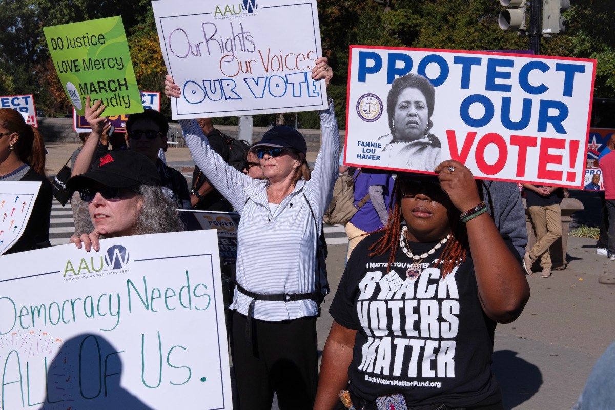people stand around holding protest signs supporting voting rights. One woman wearing a black tshirt that says "BLACK VOTERS MATTER" is holding up a sign with a black and white photo of Fannie Lou Hamer with the words "PROTECT OUR VOTE"
