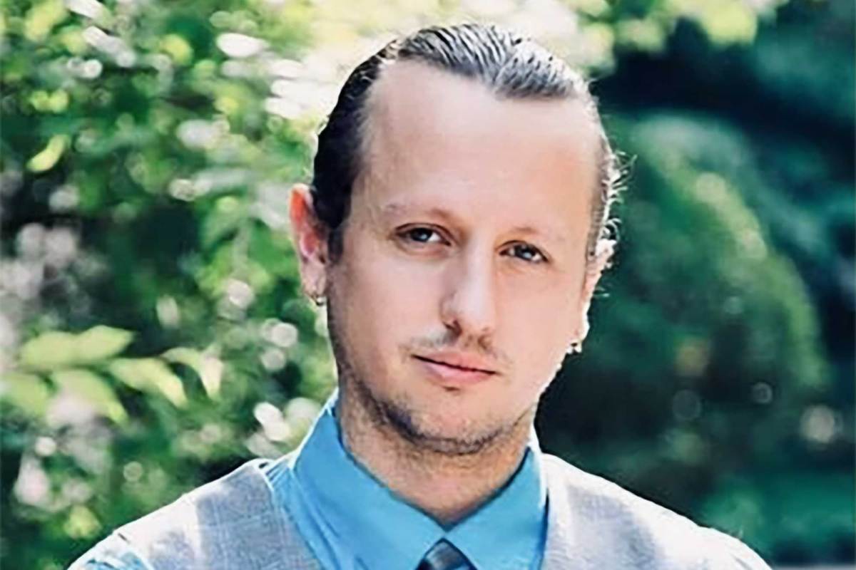 A closeup of a man in vest and tie with his hair pulled back, standing outside