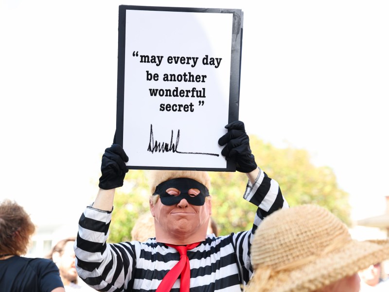 A protester dressed like Donald Trump in a cartoonish burglar costume holding up a sign that reads 'May every day be another wonderful secret' with Donald Trump's signature at the bottom