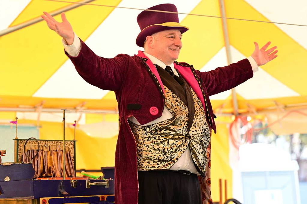 A man in a burgundy circus ringleader outfit holds his hands out wide before the flea circus