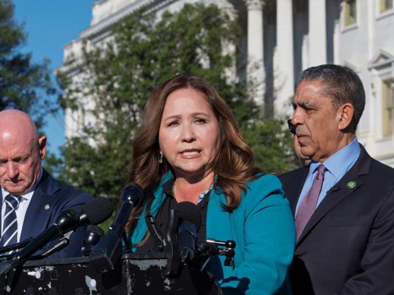 Adellita Grijalva speaking at a podium as two men on either side of her listen on.