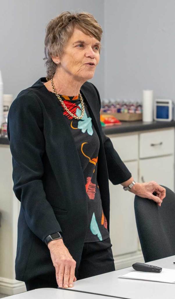 Older woman wearing a black blazer and patterned top, standing and speaking during a training session.