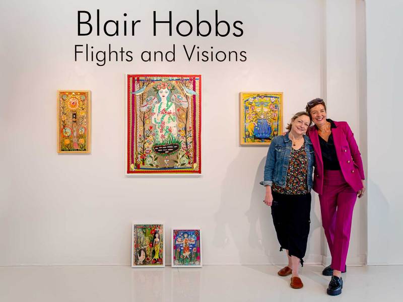 Two people stand together in front of an art exhibit. Text on the wall reads 'Blair Hobbes Flights and Visions'