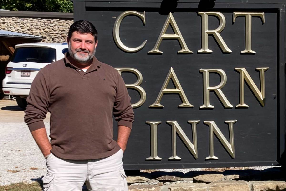 A man in a long sleeve brown top stands with hands in pockets in front of a black sign that reads "CART BARN INN" in gold letters