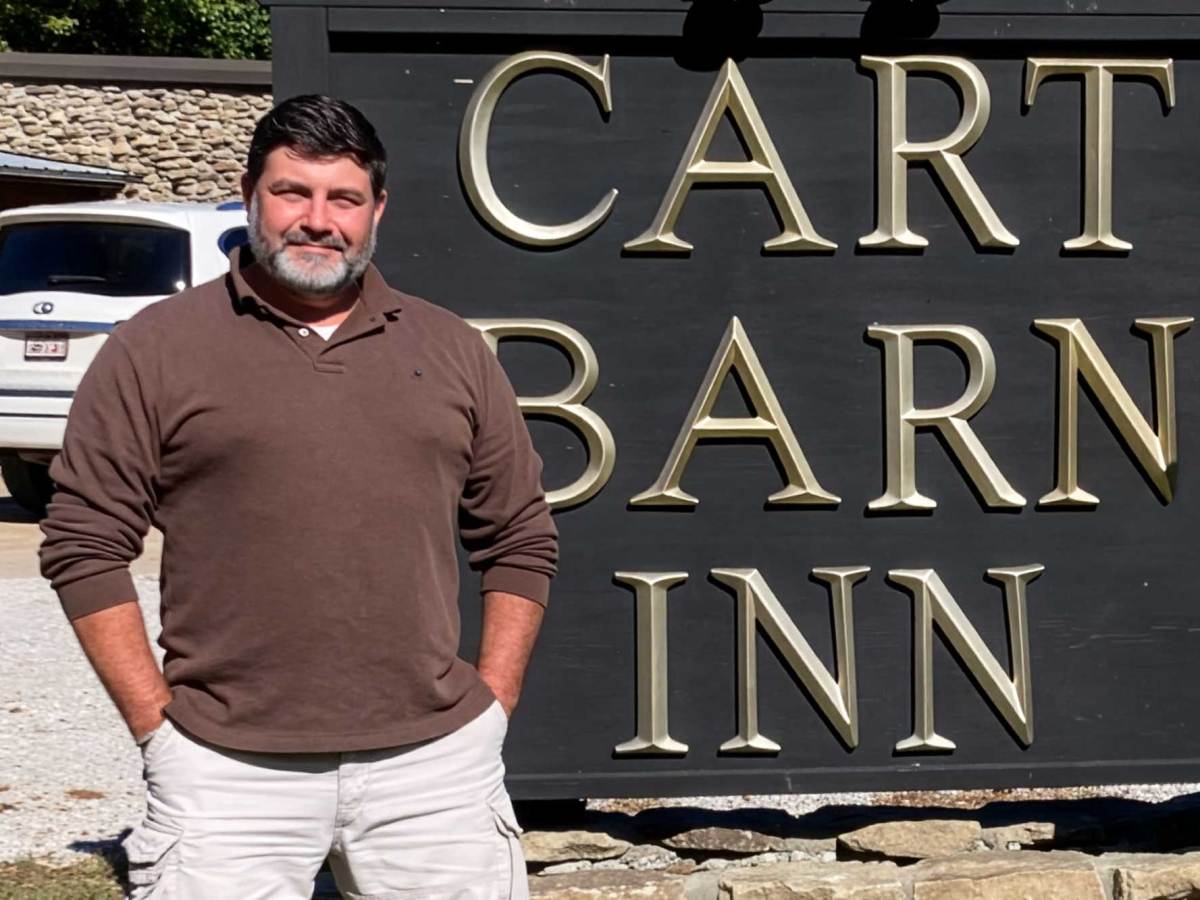 A man in a long sleeve brown top stands with hands in pockets in front of a black sign that reads "CART BARN INN" in gold letters