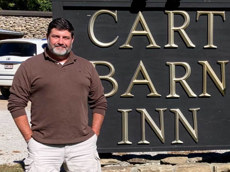 A man in a long sleeve brown top stands with hands in pockets in front of a black sign that reads "CART BARN INN" in gold letters