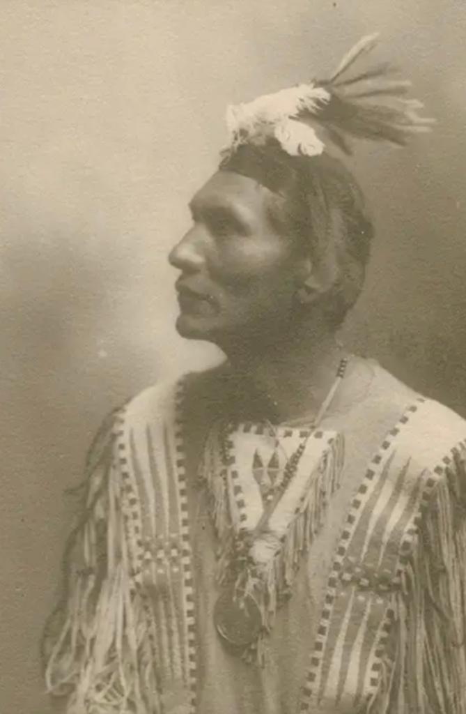 Sepia-toned portrait of man wearing a headdress and traditional Native American clothing.