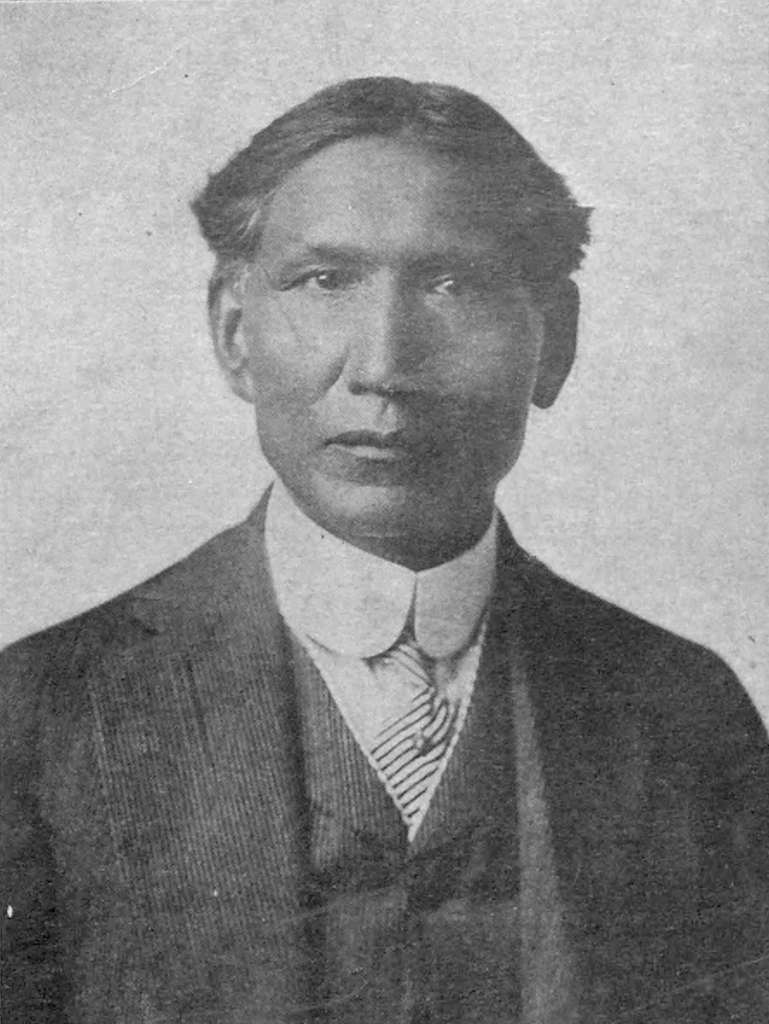 A dark-skinned, expressionless man wearing a suit and tie.