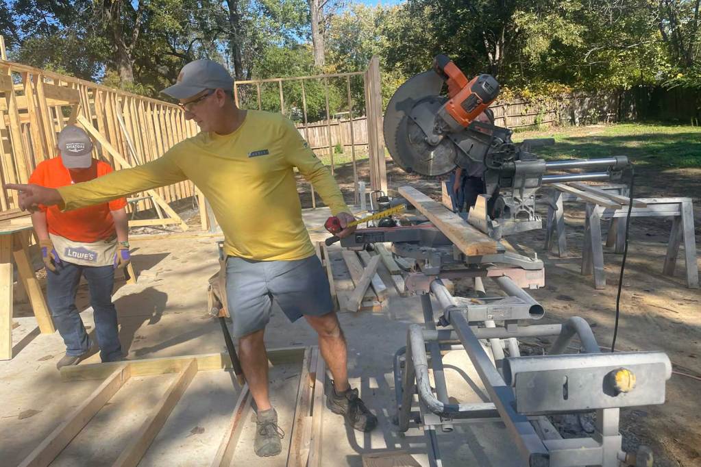 A man stands in front of a wood saw and gestures to his left at others working on a house