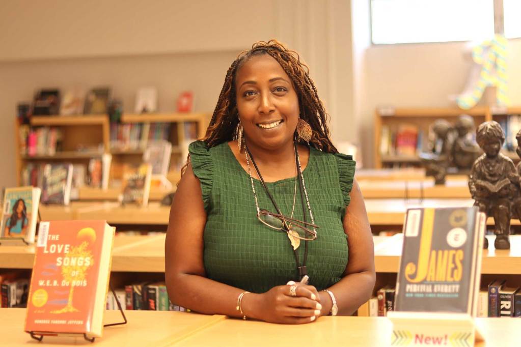A librarian dressed in green stands between shelves of books in a school library