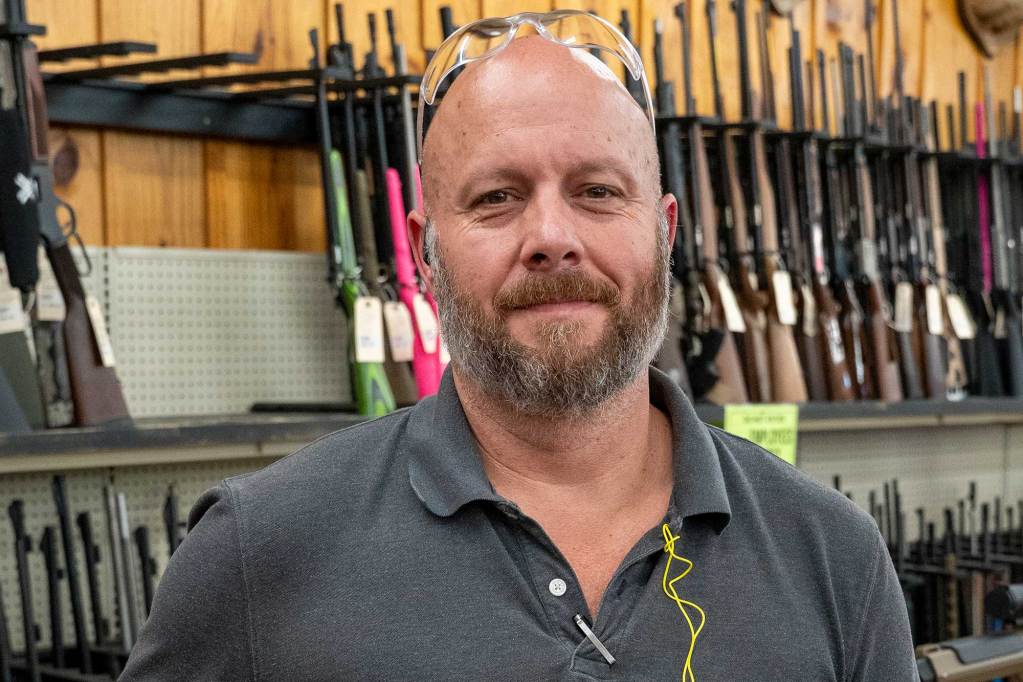 A man wearing a gray polo shirt stands in front of a gun counter lined with rifles.