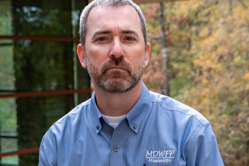 A man in a blue button-down shirt stands outdoors near a railing with trees in the background.
