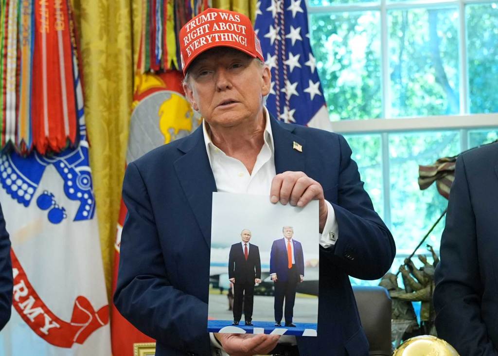 Donald Trump, wearing a red hat that reads 'Trump was right about everything' stands in the Oval office and shows off ap hoto of him and Vladimir Putin walking together