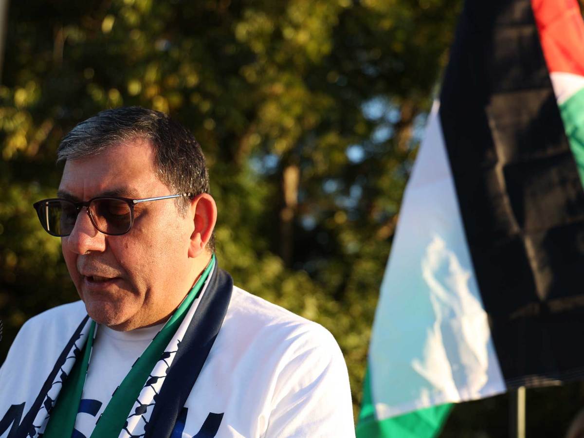 A man speaks in front of a Palestinian flag