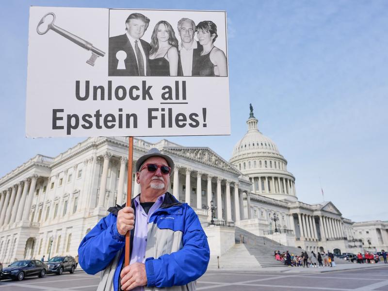 A man holding a sign in front of the U.S. Capitol building. The sign says, “Unlock all the Epstein Files!”