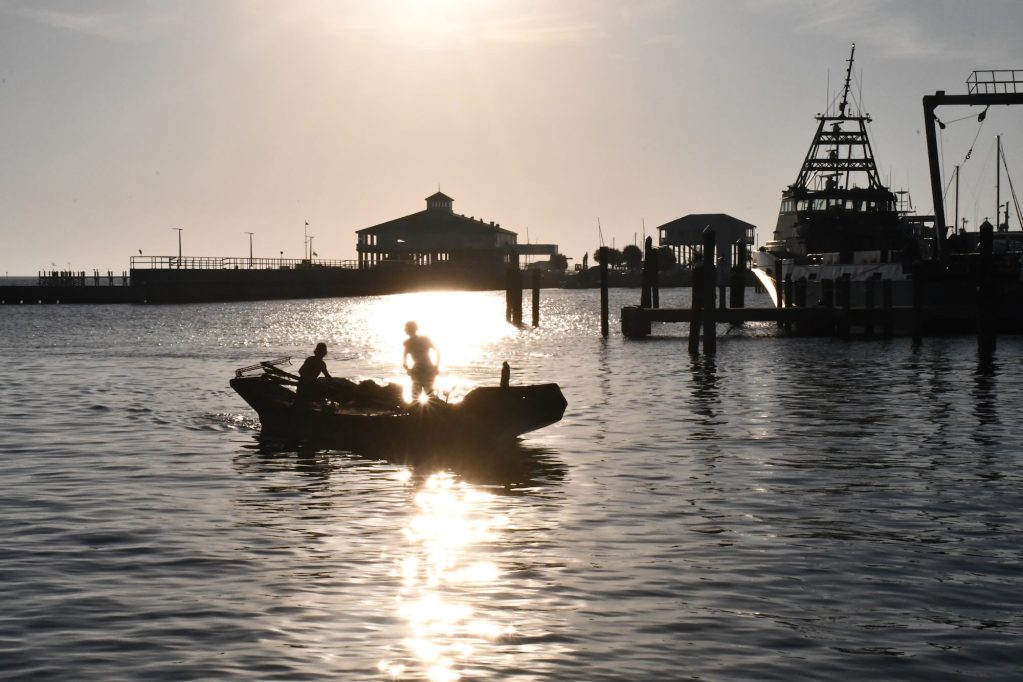 Two people stand on an oyster tonging boat silhouetted against the setting sun as they return to a harbor, with fishing vessels and dock buildings visible in the background.