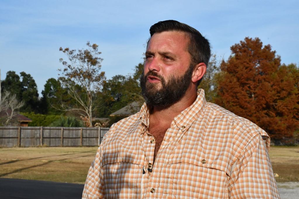 A man in a orange and white button up stands in the sun with a fence and trees behind him.