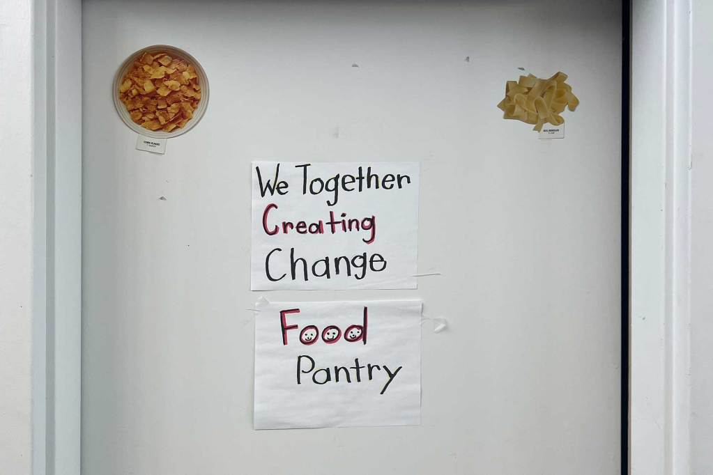 A door with a hand written sign that reads 'We Together Creating Change Food Pantry'. Tiny photos of food are taped above.