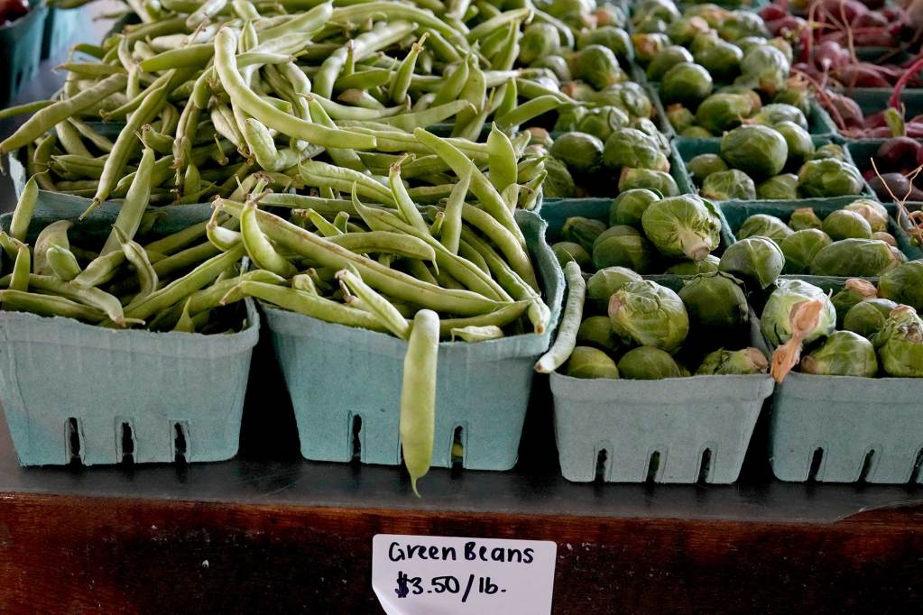 Cartons of Green beans and brussel sprouts sit on a table with a sign that reads ‘Green Beans $3.50/lb’