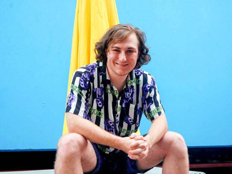 A man in a colorful striped shirt sits in front of a blue wall