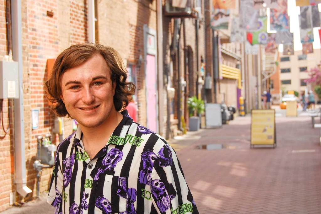 A man in a colorful striped shirt stands outside in a brick lined alley