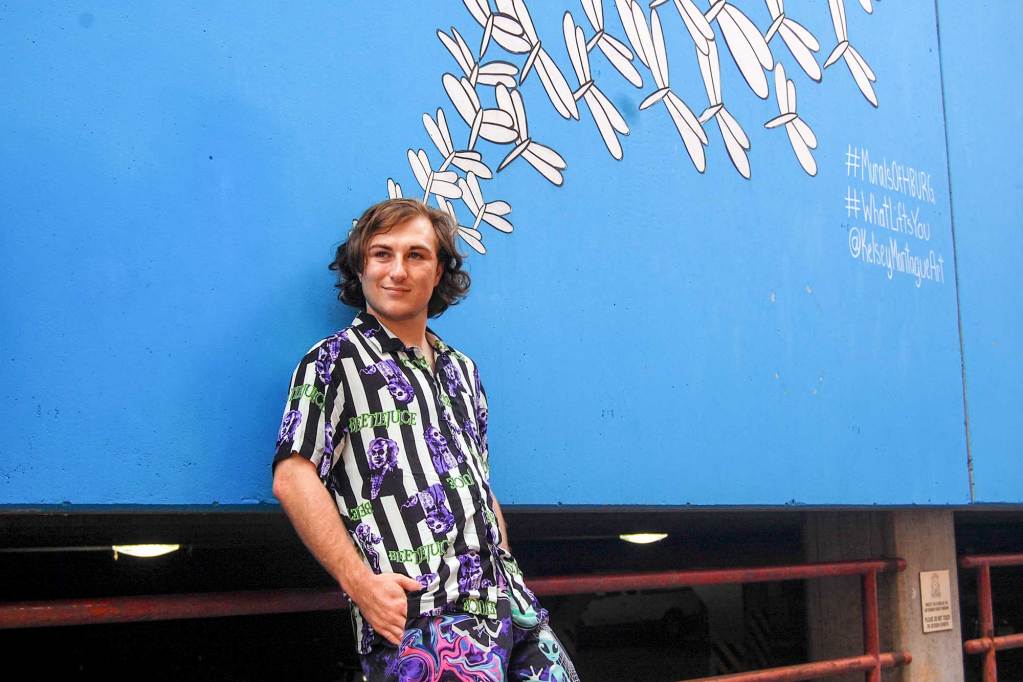 A man in a colorful striped shirt leans against a blue wall with a simple dragonfly mural