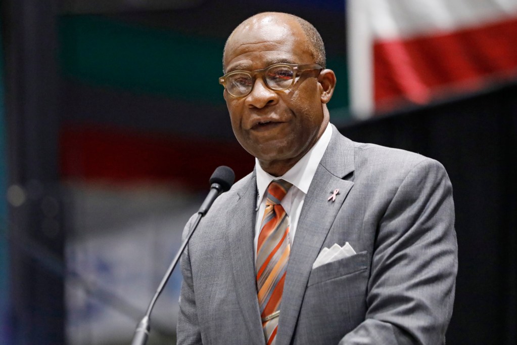 A Black man in a gray suit with glasses standing at a podium