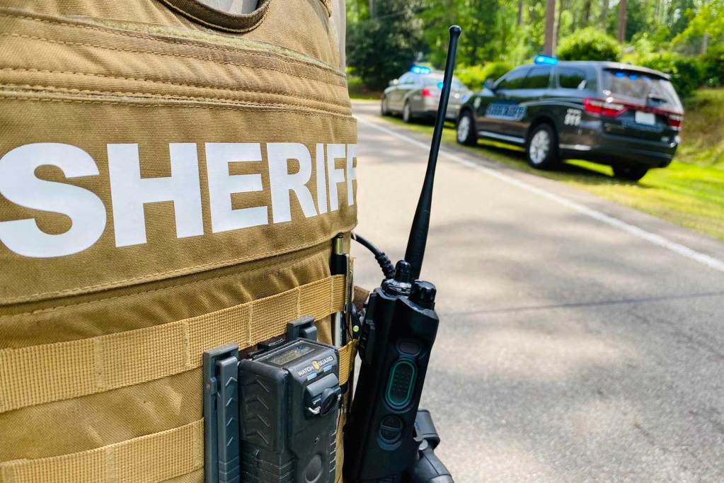 A closeup view of a Sheriff's brown vest with walkie talkie and other things attached at the belt. A sheriff's car can be seen on the side of the road in the background