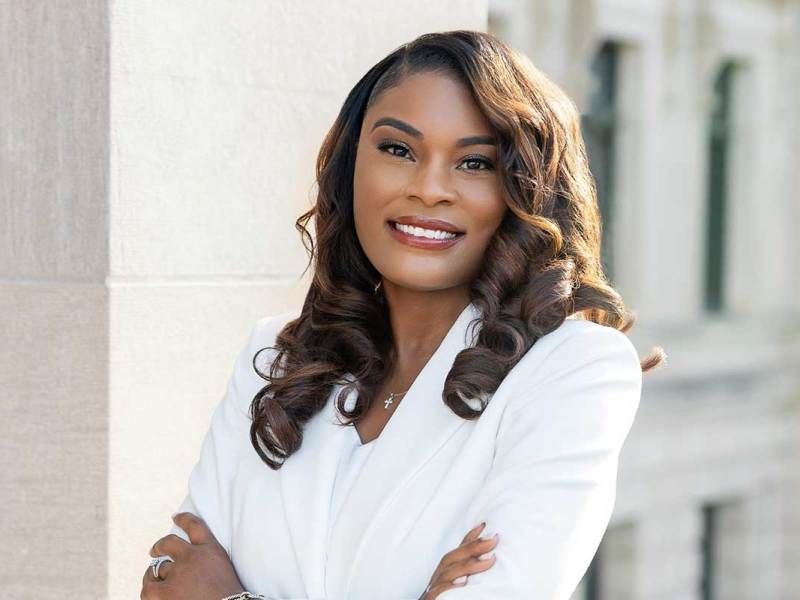 A woman wearing a white blazer stands in front of a courthouse with her arms crossed.