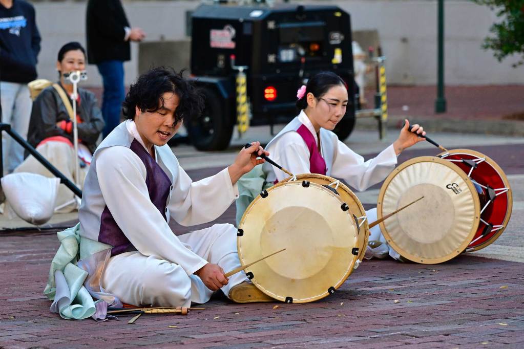 Two Korean performers sit on the ground and play drums as a woman in traditional Korean dress sits behind and listens on