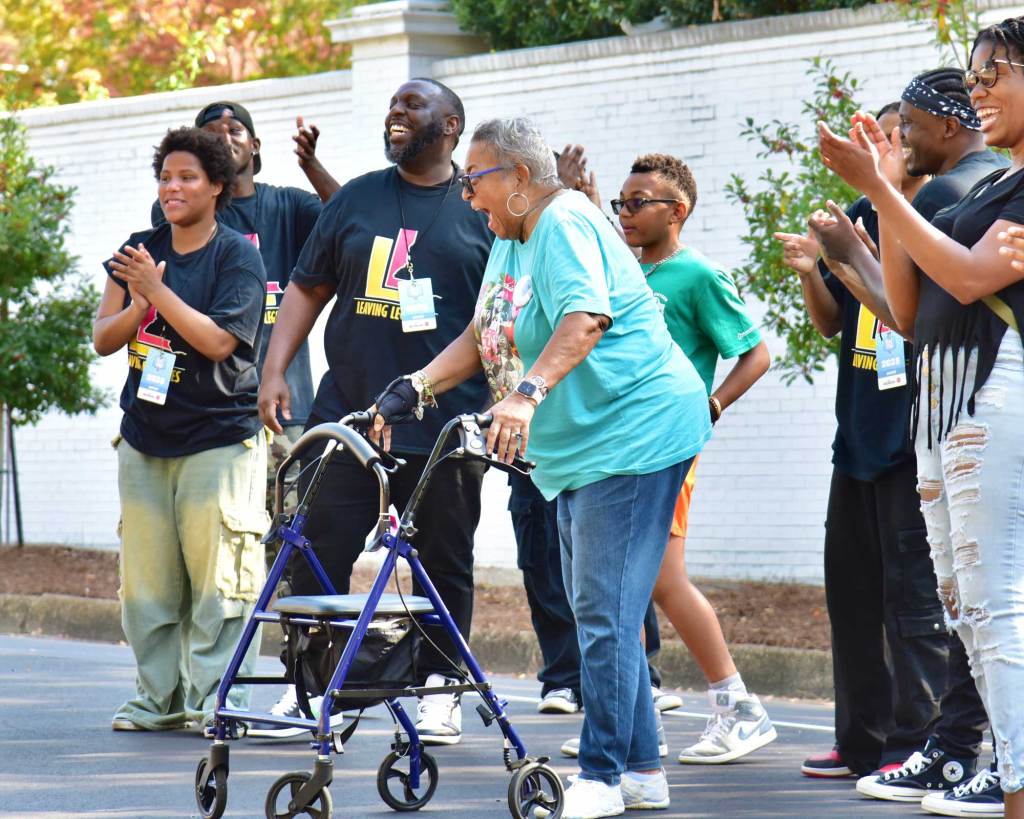 Men in matching black shirts stand around an older woman with a walker, everyone clapping
