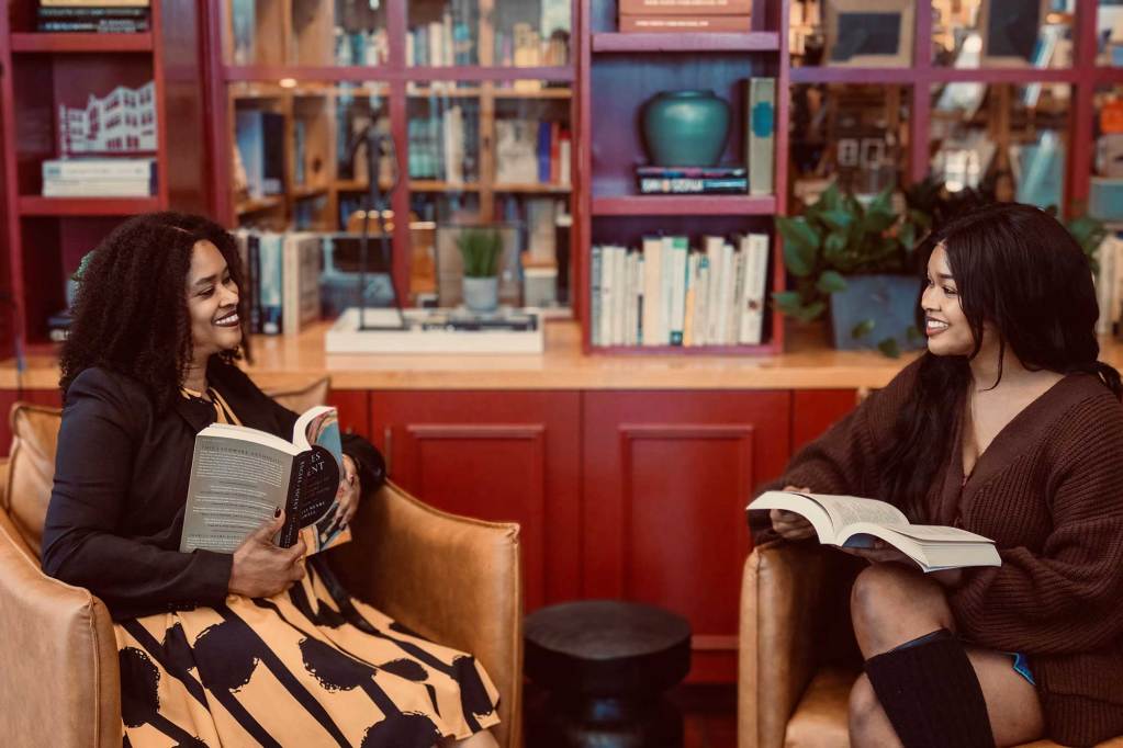 Two women sit in chairs next to each other and smile as they both hold open books in their laps