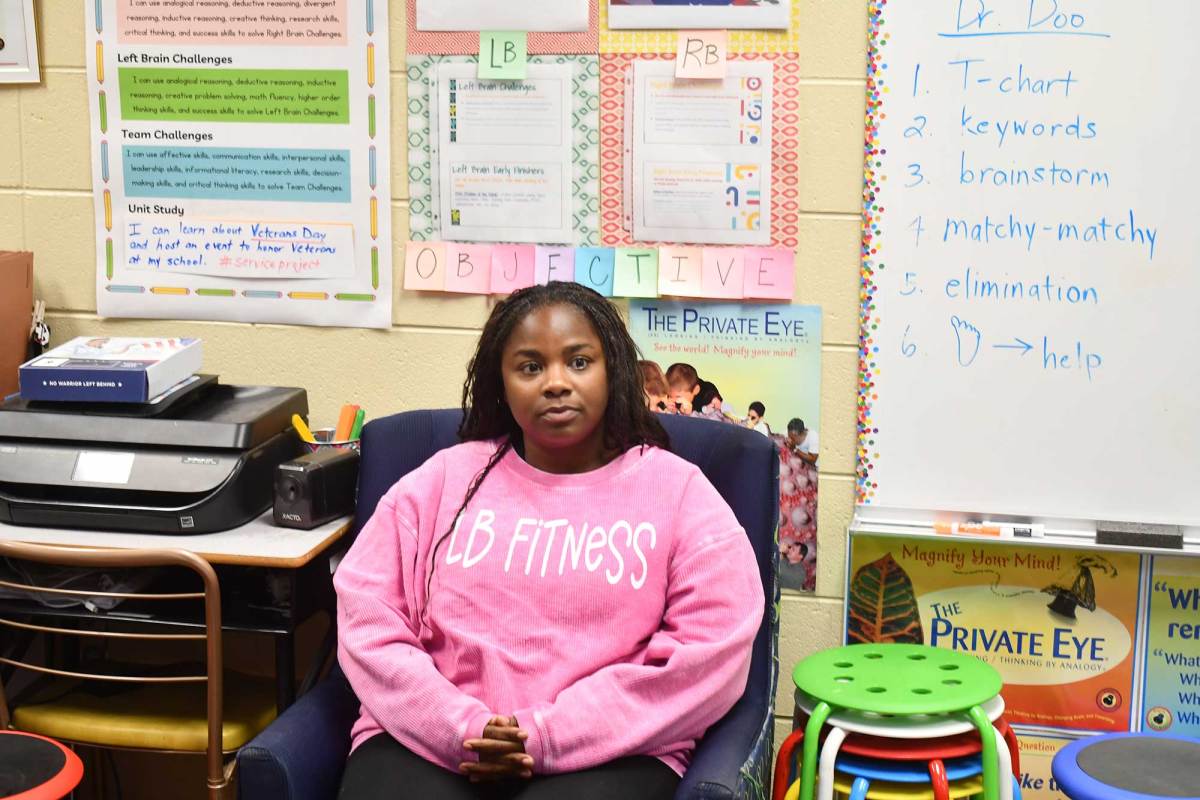 A woman in a pink sweatshirt sits in a blue suede chair inside a classroom surrounded by colorful posters and a whiteboard.