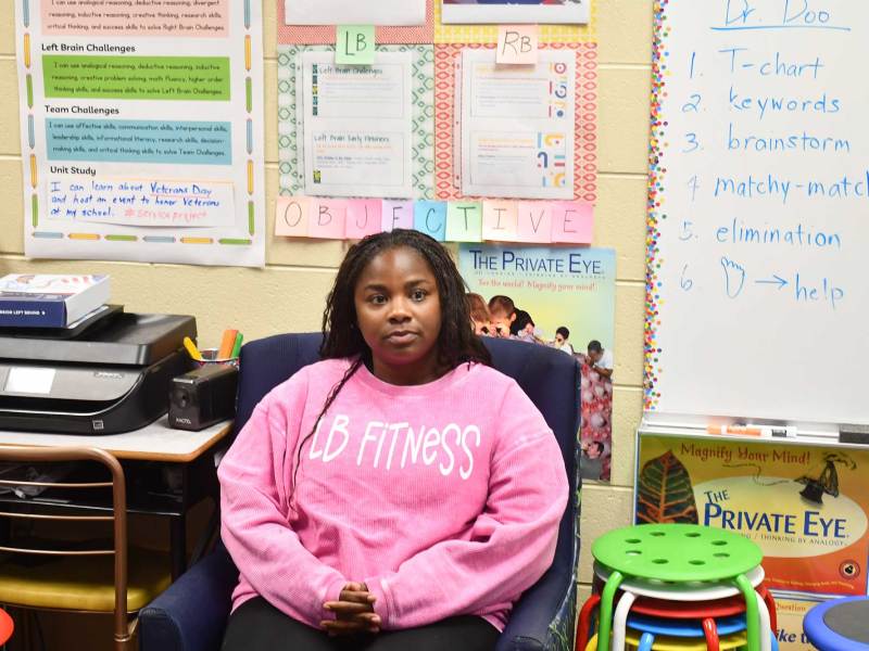 A woman in a pink sweatshirt sits in a blue suede chair inside a classroom surrounded by colorful posters and a whiteboard.
