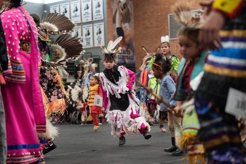 A room full of Lumbee Tribe members in traditional dress perform, a young member dancing in the center