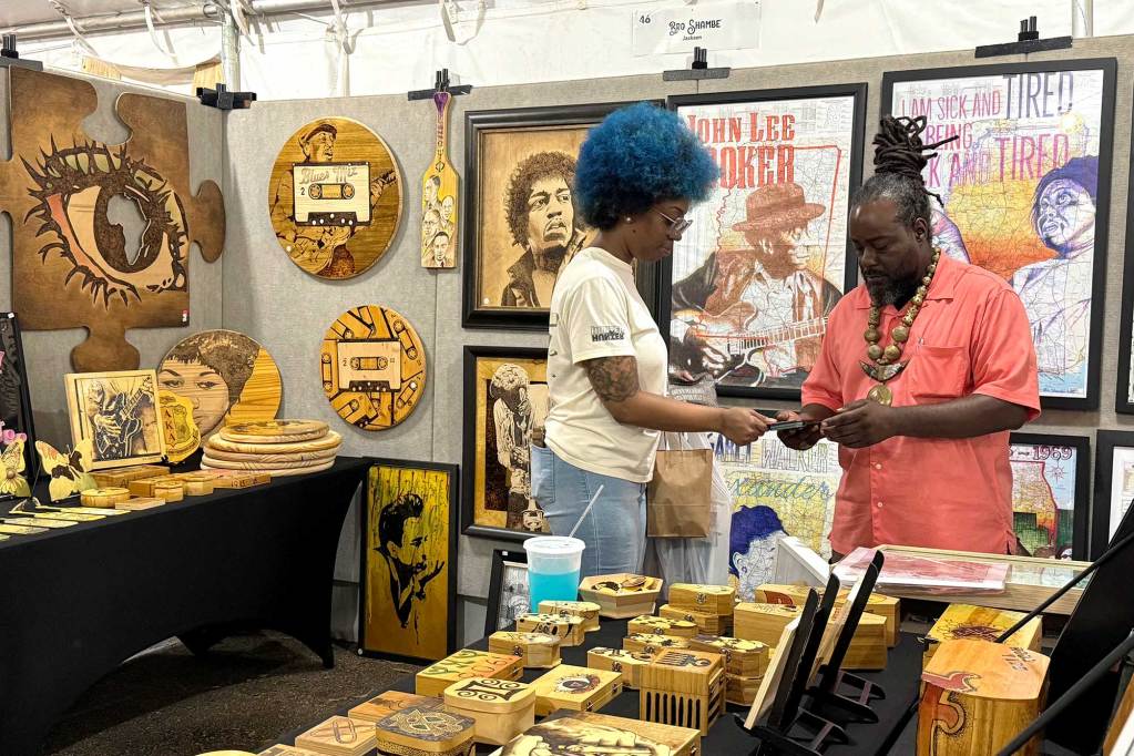 A man and a woman stand inside an art booth filled with engraved wood designs