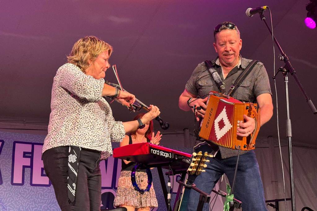 A woman plays a violin as a man beside her plays an accordion, and another woman is visible with a keyboard behind them both