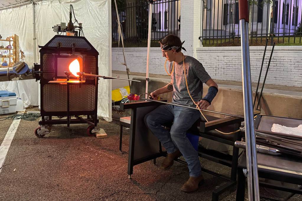 A man blows glass in a hot kiln outside at a vendor's booth