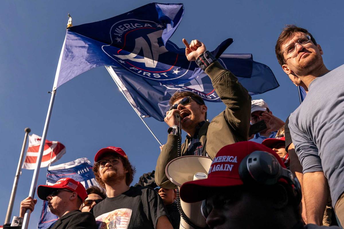 Nick Fuentes speaking into a hand-mic. Other men surround him with flags and hats that say “Make America Great Again.”