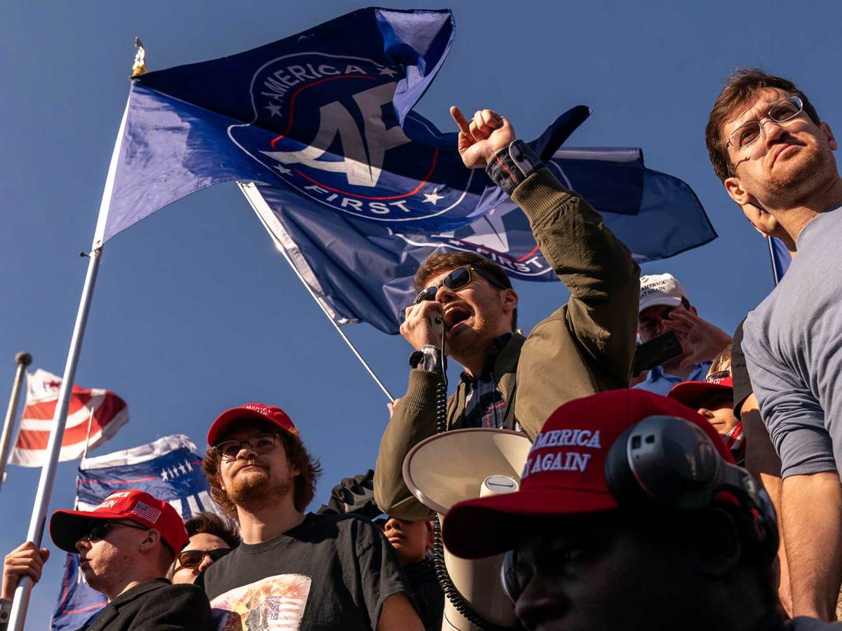 Nick Fuentes speaking into a hand-mic. Other men surround him with flags and hats that say “Make America Great Again.”