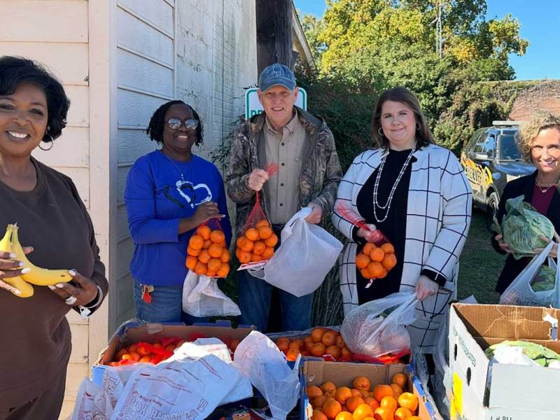 Five people stand outside holding produce behind boxes containing more fruits and vegetables