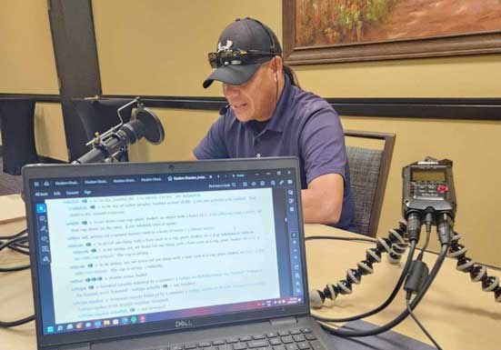 A man speaks into a microphone set on a table, and text is seen on a laptop