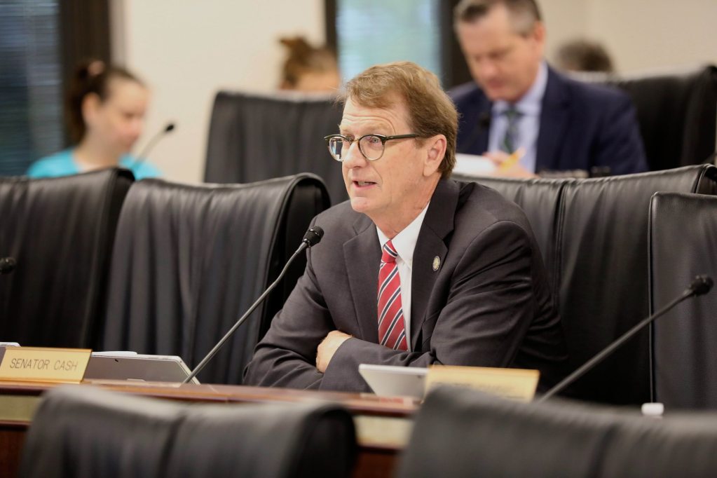 a white man with glasses sits at a committee desk in front of a microphone and a sign that says "SENATOR CASH"