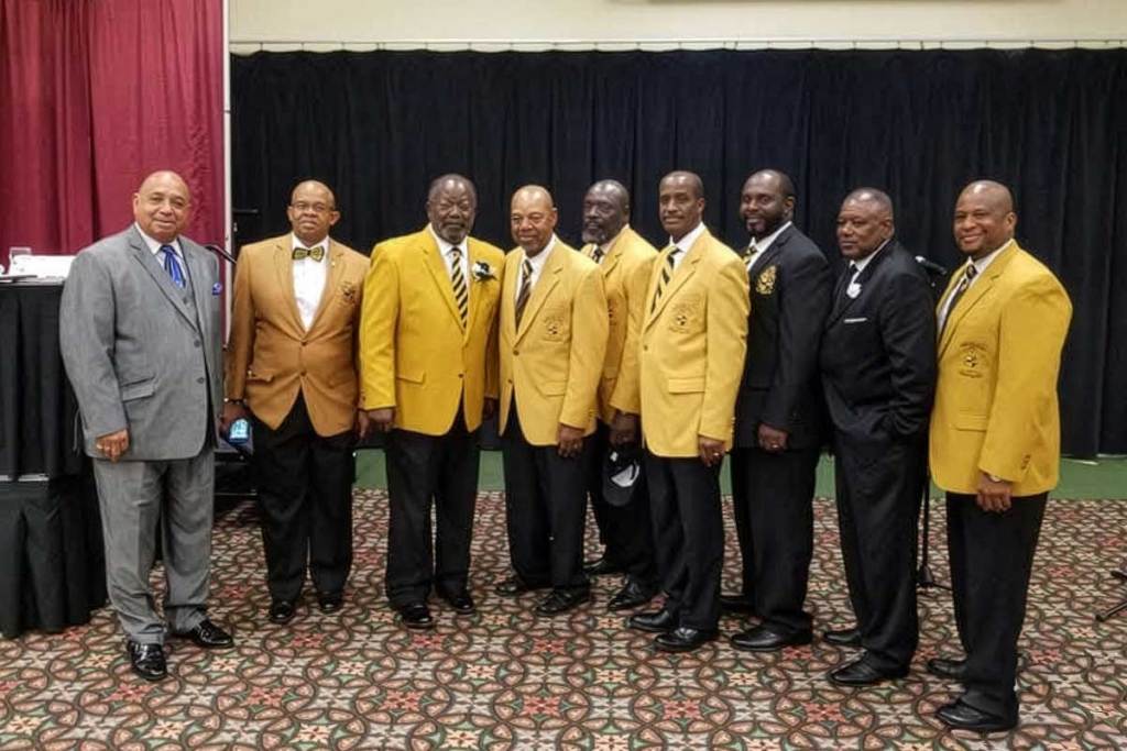 A group of men, most wearing yellow jackets with matching yellow and black striped ties, stand together in a line